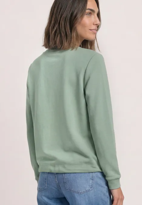 Frau von hinten in grünem Sweatshirt und blauer Jeans vor neutralem Hintergrund.