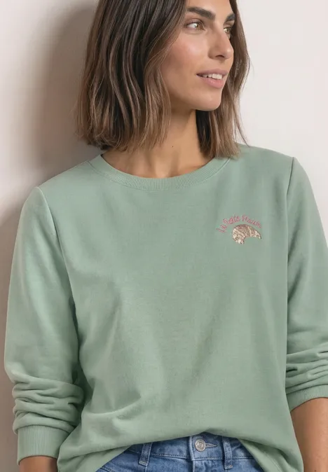 Frau trägt mintgrünes Sweatshirt mit dezentem Motiv und Jeans, blickt seitlich.