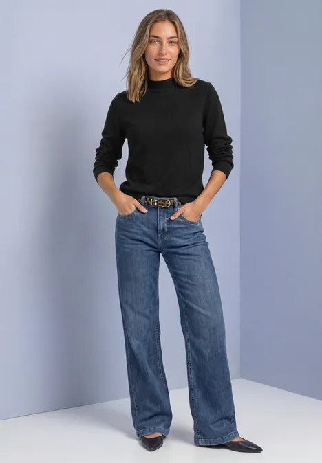 Frau mit schwarzem Pullover, weitem blauen Jeans, Gürtel und spitzen Schuhen vor neutralem Hintergrund.