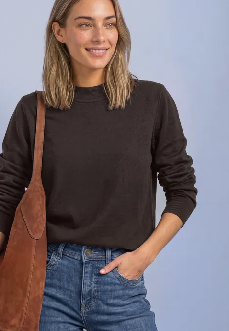 Frau mit dunkelbraunem Pullover, Jeans und brauner Ledertasche lächelt vor hellem Hintergrund.