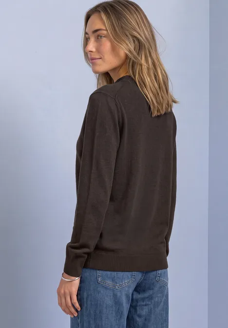 Frau in dunklem Pullover und blauen Jeans steht seitlich vor hellem Hintergrund.