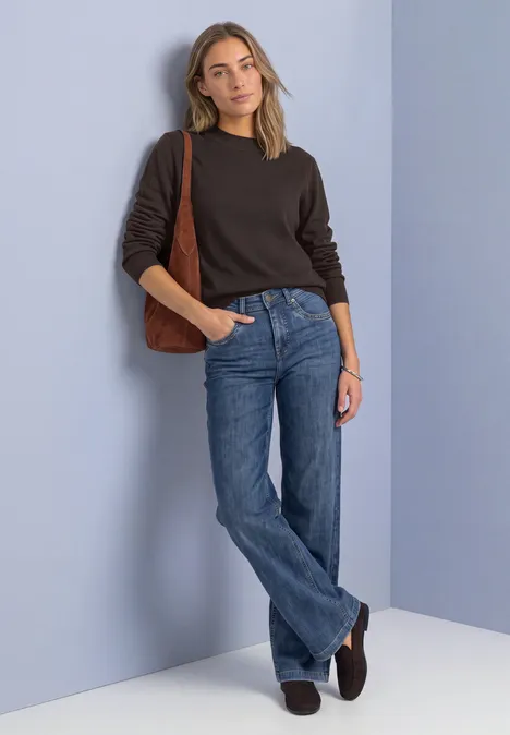Eine Frau lehnt entspannt an lilafarbener Wand, trägt schwarzen Pullover, blaue Jeans und braune Schultertasche.