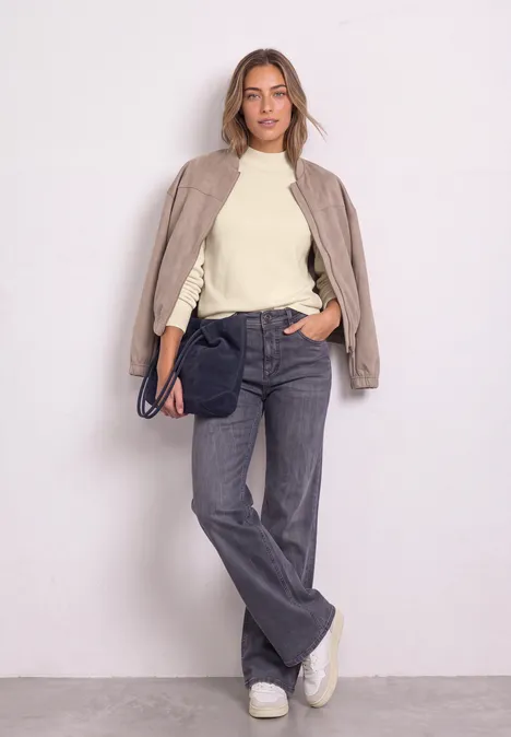Eine Frau in beige Bomberjacke, cremefarbenem Pullover, grauen Jeans und weißen Sneakern lehnt an weißer Wand.
