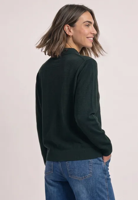 Eine junge Frau im dunkelgrünen Pullover und Jeans lächelt, während sie über die Schulter schaut.