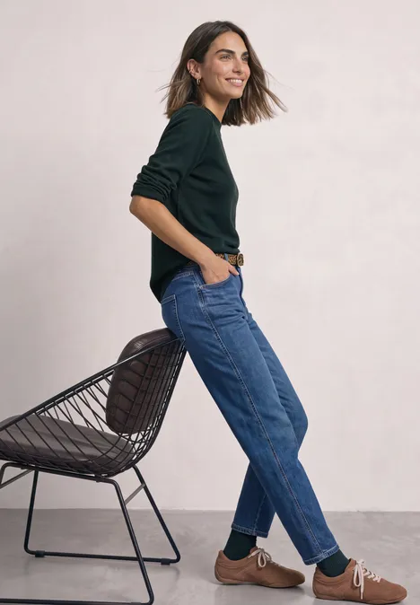 Eine Frau in schwarzem Pullover und blauen Jeans lehnt lächelnd an einem Stuhl vor hellgrauem Hintergrund.