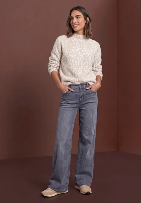 Pull en laine fine à motif jacquard clay sand Frau im hellen Strickpullover und weiten Jeans vor braunem Hintergrund.