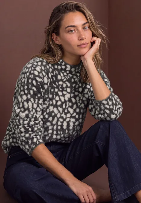 Pull en laine fine à motif jacquard black coffee Eine Frau sitzt entspannt in Jeans und grauem, weiß gepunktetem Pullover vor schlichtem Hintergrund.