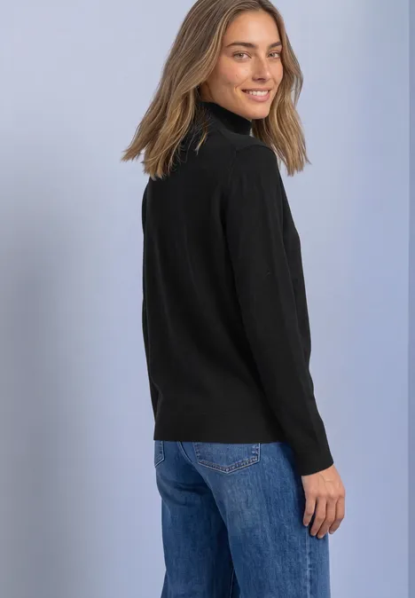 Pullover mit Turtle Neck und Knopfdetails Black Junge Frau mit schwarzem Rollkragenpullover und blauen Jeans, schaut lächelnd über die Schulter vor hellem Hintergrund.