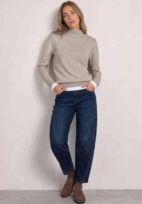 Frau in beige Rollkragenpullover, weißem Shirt und dunkelblauen Jeans steht lässig vor hellem Hintergrund.