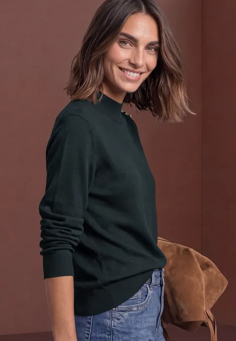 Pullover mit Turtle Neck und Knopfdetails stormy green Lächelnde Frau in grünem Pullover und Jeans mit brauner Tasche vor rotem Hintergrund.