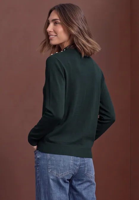 Pullover mit Turtle Neck und Knopfdetails stormy green Frau im dunkelgrünen Pullover und Jeans, im Profil stehend, vor braunem Hintergrund.