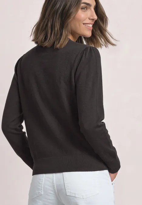 Frau in schwarzem Pullover und weißer Jeans, von hinten mit Blick über die Schulter, lächelnd.
