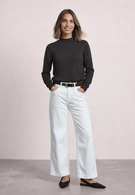 Frau in weitem weißen Jeans, schwarzem Oberteil, Gürtel und Ballerinas, lächelnd vor neutralem Hintergrund.