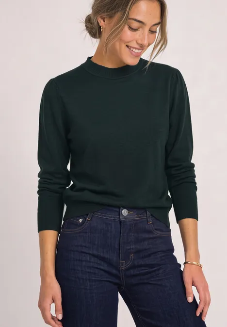 Vrouw in donkergroene trui en jeans lacht en kijkt naar beneden.
