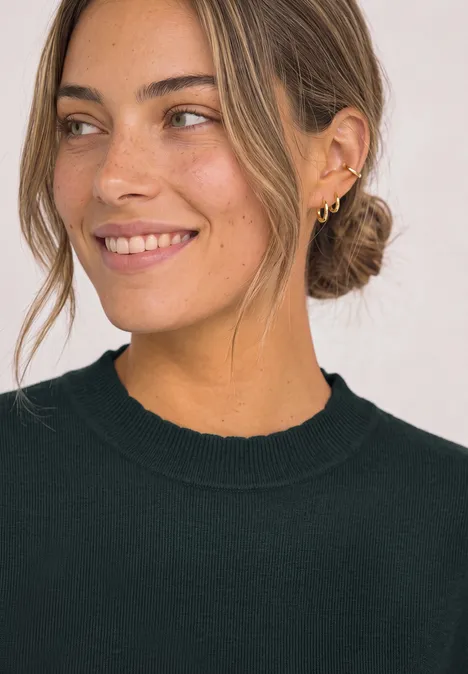 Vrouw met bruin knotje, meerdere gouden oorbellen en zwarte trui, glimlachend.
