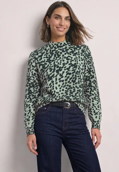 Een glimlachende vrouw draagt een groen-zwart dierenprintblouse met lange mouwen en een donkere spijkerbroek.