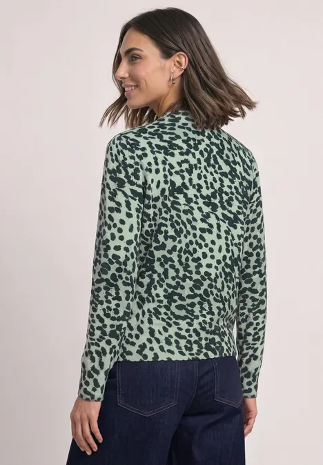 Vrouw in groen-zwart gevlekt shirt met lange mouwen en spijkerbroek, driekwart achteraanzicht, ze lacht.