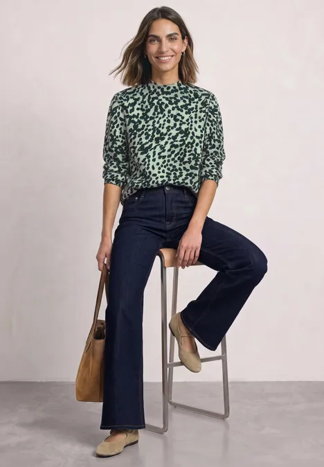 Vrouw in groen dessinblouse en donker spijkerbroek zit ontspannen op een hoge kruk, glimlachend.