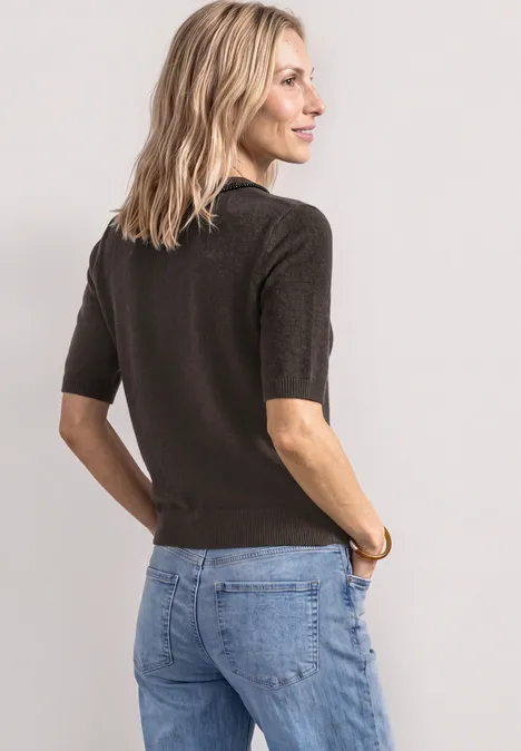 Eine Frau in schlichter, dunkelbrauner Strickbluse mit halblangen Ärmeln und blauen Jeans vor hellem Hintergrund.