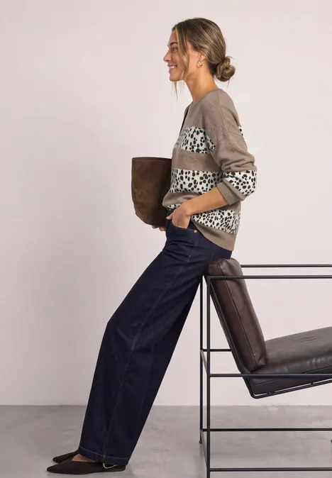 Femme en gilet à motif et jean large appuyée sur une chaise, tenant une veste.
