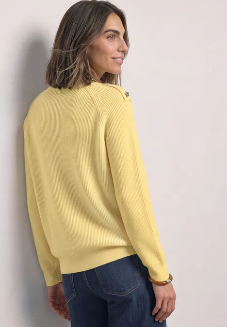 Frau in gelbem Pullover und blauen Jeans, steht seitlich mit Blick über die Schulter und lächelt.