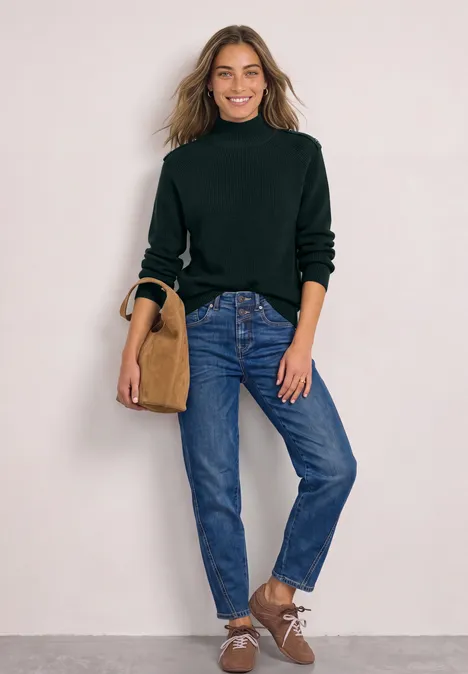 Pullover mit Turtle Neck und Knopfdetails stormy green Frau in dunkelgrünem Rollkragenpullover, blauen Jeans und braunen Schuhen mit Tasche vor hellem Hintergrund.