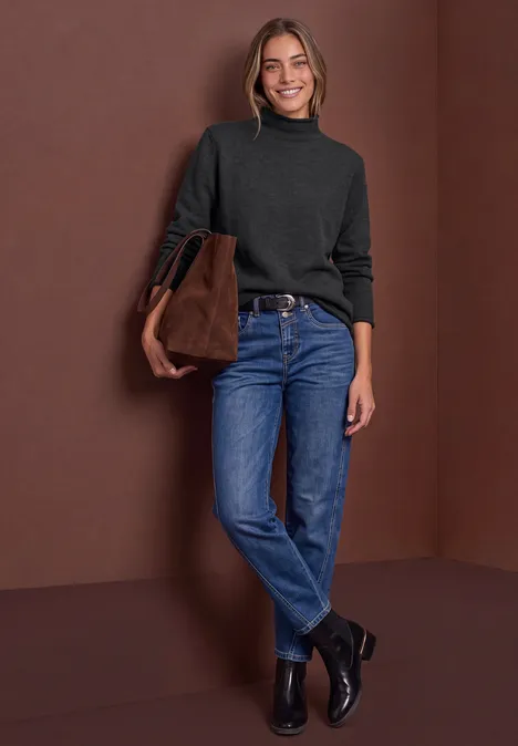 Pullover mit Turtle Neck und Rollkanten armor grey mel. Eine Frau in Jeans, schwarzem Rollkragenpullover und Stiefeletten, trägt eine braune Tasche vor braunem Hintergrund.