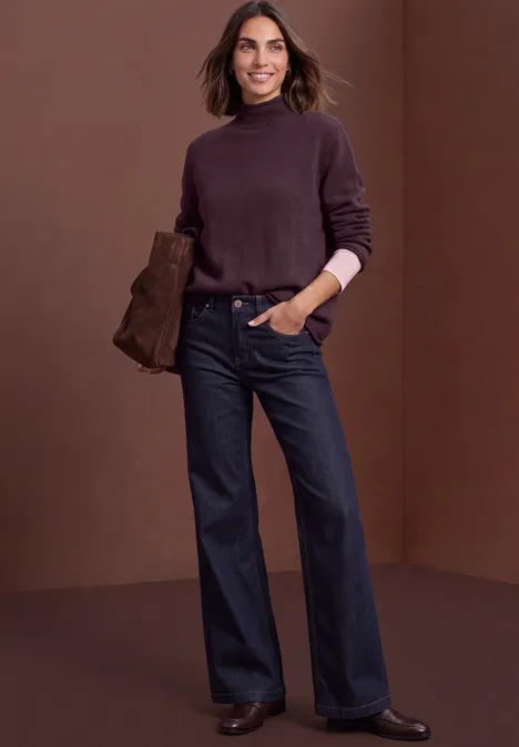 Pull à col roulé et bords roulés berry plum mel. Frau in dunkelviolettem Rollkragenpullover und weiten Jeans hält eine Clutch vor braunem Hintergrund.