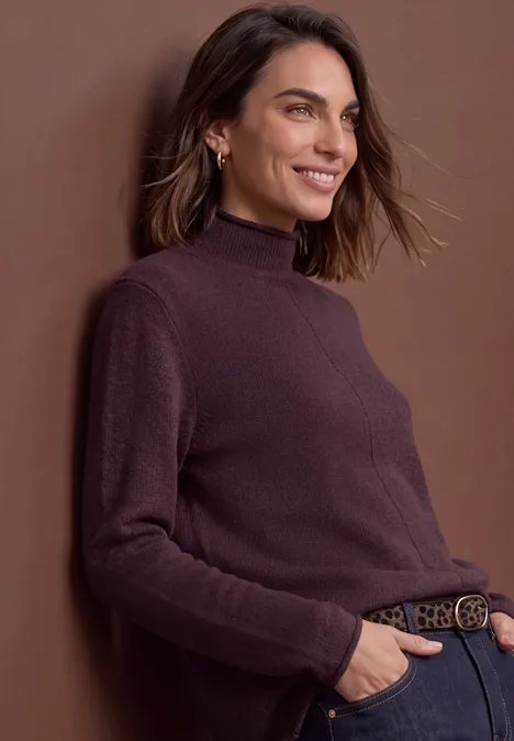 Pull à col roulé et bords roulés berry plum mel. Eine lächelnde Frau, die in braunem Rollkragenpullover und Jeans an eine Wand gelehnt steht.