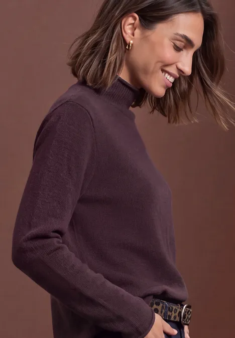 Pull à col roulé et bords roulés berry plum mel. Frau in violettem Rollkragenpullover, lächelnd in Seitenansicht vor schlichtem braunem Hintergrund.