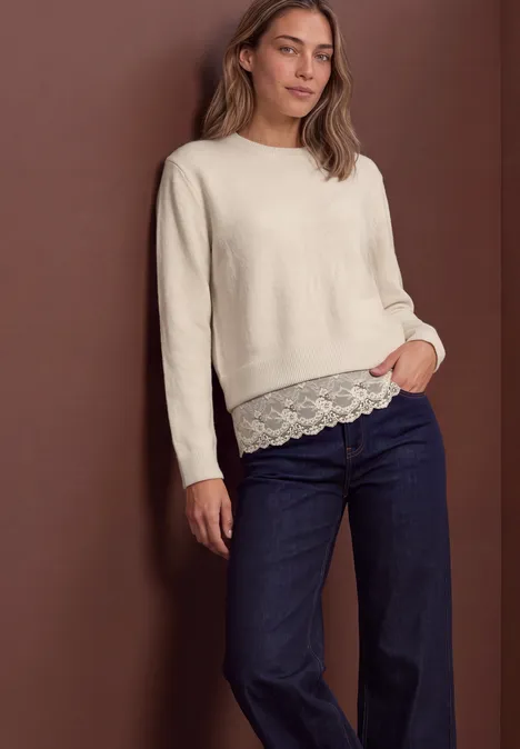 Pullover mit Rundhals und Spitzenkante pure beige Frau trägt cremefarbenen Pullover mit Spitzenbesatz unten und dunkle Jeans, steht vor braunem Hintergrund.
