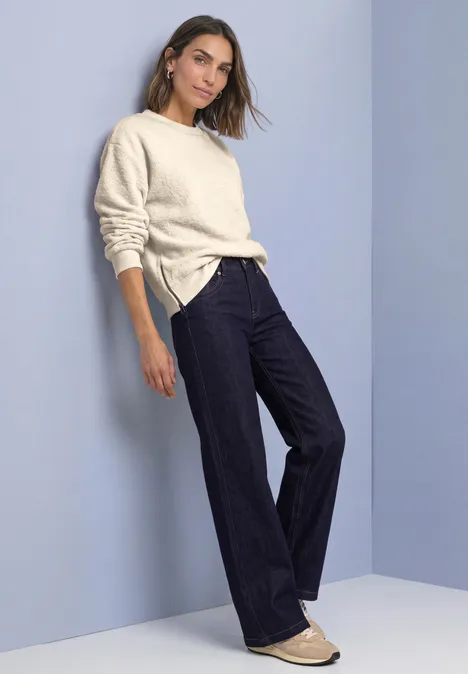 Teddy-Sweatshirt mit Zipperdetails pure beige Frau lehnt an Wand, trägt cremefarbenen Pullover, dunkle Schlagjeans und helle Ballerinas vor schlichtem Hintergrund.