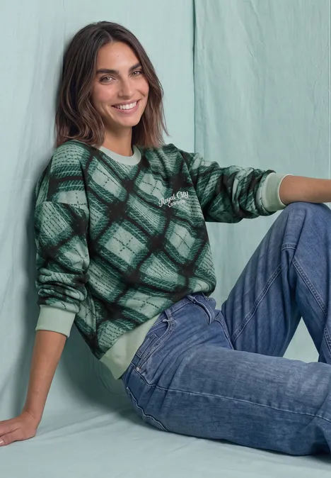 Sweat-shirt à col rond et motif à carreaux stormy green Une femme souriante posant devant un mur vert pastel, vêtue d’un pull vert à carreaux et d’un jean.