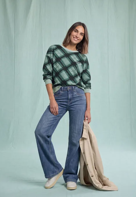 Sweat-shirt à col rond et motif à carreaux stormy green Femme en haut à carreaux verts, jean flare bleu, baskets blanches, manteau beige à la main devant un fond vert.