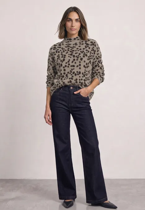 Frau in weitem dunkelblauen Jeans, leo-gemustertem Pullover, vor neutralem Hintergrund stehend.