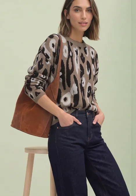 Frau mit Animal-Print-Pullover, dunklen Jeans und brauner Tasche lehnt lässig an einem Hocker.
