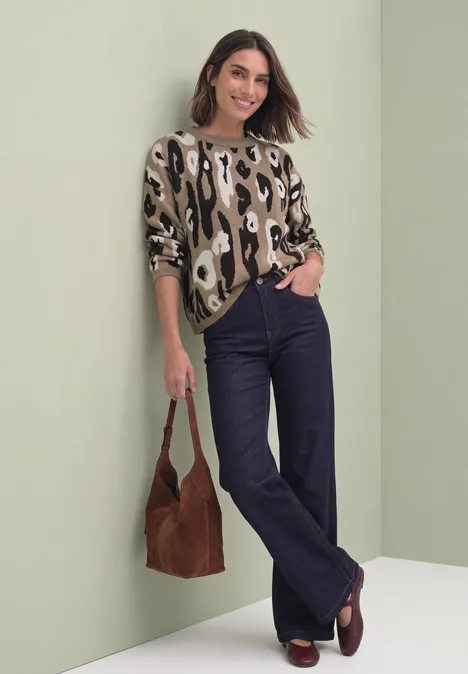 Frau in Leopardenmuster-Pullover, weiten Jeans und braunen Loafers lehnt lächelnd an hellgrüner Wand.