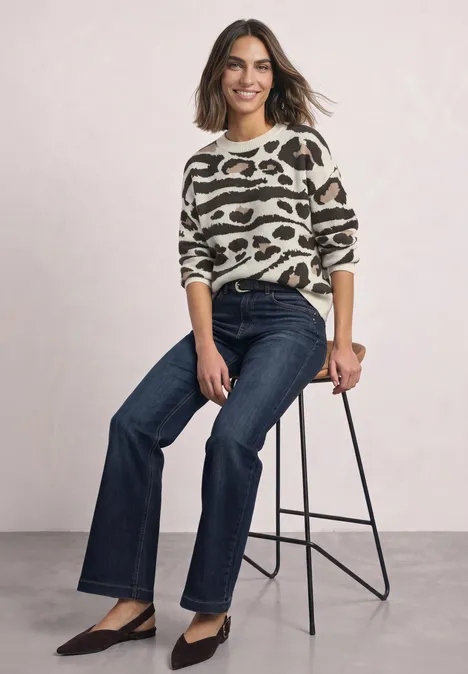 Femme assise sur un tabouret, en pull imprimé animal, jean bleu foncé et escarpins noirs, souriante.