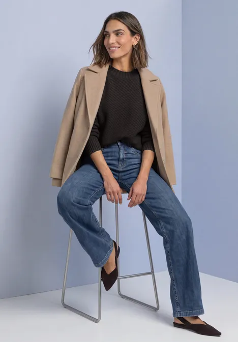 Langarm Pullover mit Turtle Neck und Struktur black coffee Frau sitzt lässig auf Hocker, trägt beigen Blazer über schwarzen Top, Jeans und spitze Schuhe.