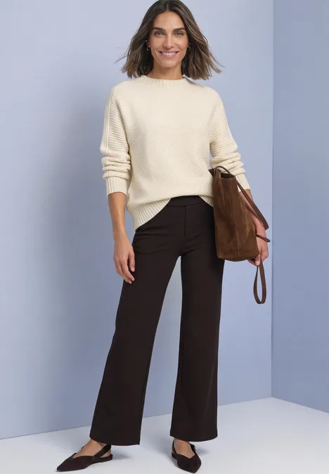 Frau in cremefarbenem Pullover, dunkelbrauner Hose und brauner Tasche vor schlichtem Hintergrund.