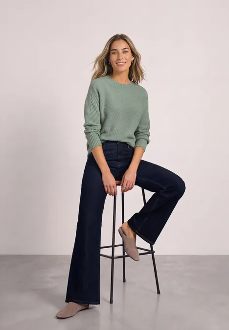 Langarm Pullover mit Turtle Neck und Struktur eucalyptus green mel. Frau in grünem Pullover und dunklen Schlagjeans sitzt lässig auf einem Hocker vor neutralem Hintergrund.
