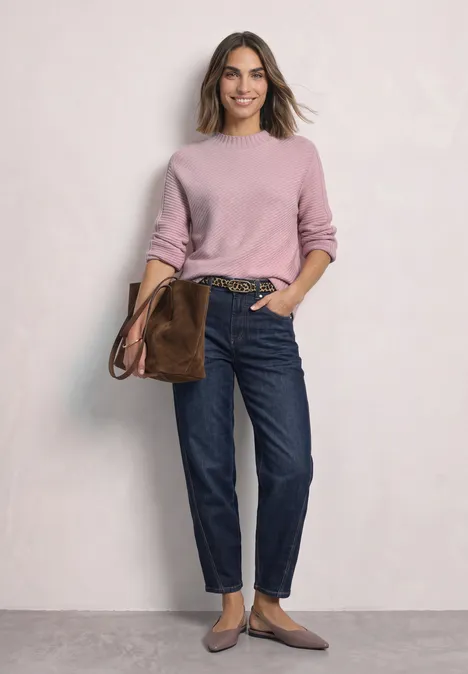 Frau in rosafarbenem Pullover, dunklen Jeans, brauner Tasche und flachen Schuhen vor hellem Hintergrund.
