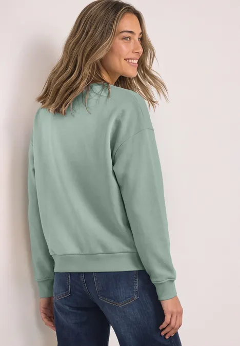 Loose Fit Sweatshirt mit Patch Detail eucalyptus green Eine junge Frau in hellgrünem Pullover schaut lächelnd über die Schulter.