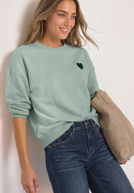 Loose Fit Sweatshirt mit Patch Detail eucalyptus green Frau in grünem Pullover mit Herz-Logo und Jeans, hält beige Jacke, lächelt vor hellem Hintergrund.