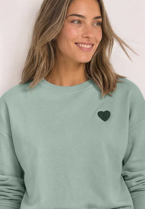 Loose Fit Sweatshirt mit Patch Detail eucalyptus green Frau lächelt in hellgrünem Sweatshirt mit kleinem schwarzem Herz-Logo auf der Brust.
