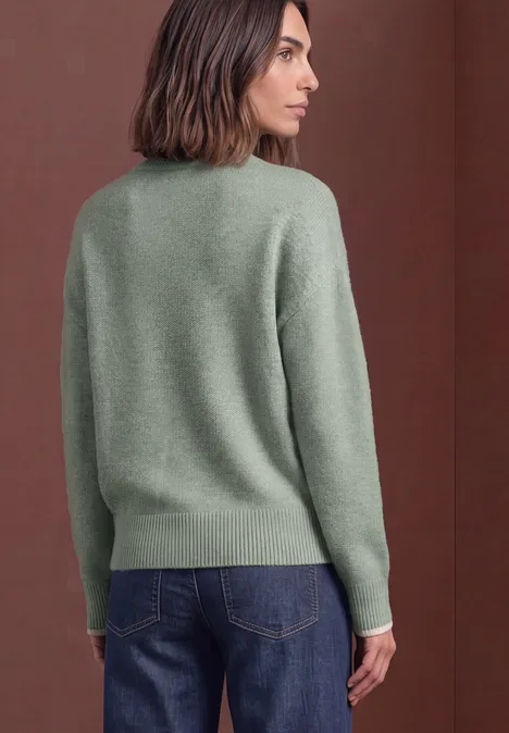 Frau von hinten in grünem Pullover und blauen Jeans vor braunem Hintergrund.