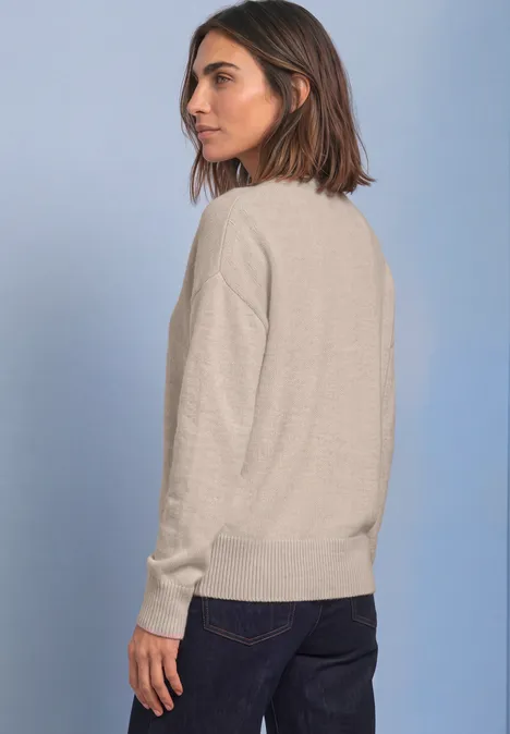 Pull uni à col rond clay sand mel. Junge Frau in hellbeigem Rundhalspullover und dunklen Hosen, seitlich stehend vor hellgrauem Hintergrund.