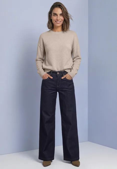 Pull uni à col rond clay sand mel. Frau mit beigem Pullover und weit geschnittenen dunklen Jeans, Hände in den Taschen, vor hellblauem Hintergrund.