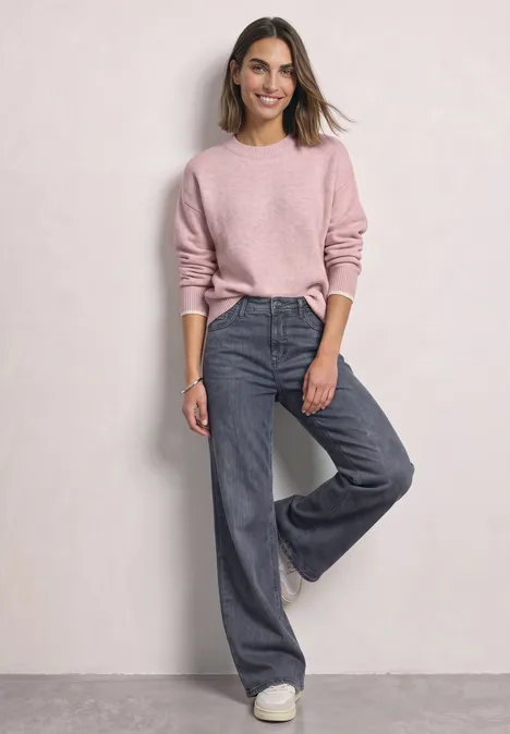 Pull uni à col rond quiet rose mel. Frau in rosa Pullover und weiten Jeans vor neutralem Hintergrund.