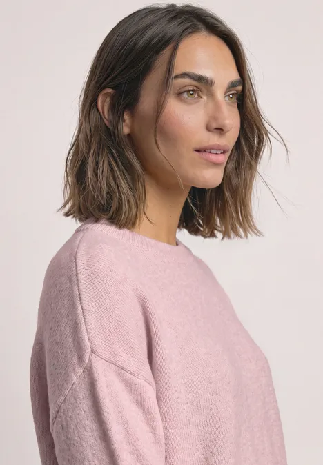 Pull uni à col rond quiet rose mel. Porträt einer Frau mit braunen Haaren in rosa Pullover vor hellem Hintergrund, Blick seitlich rechts.